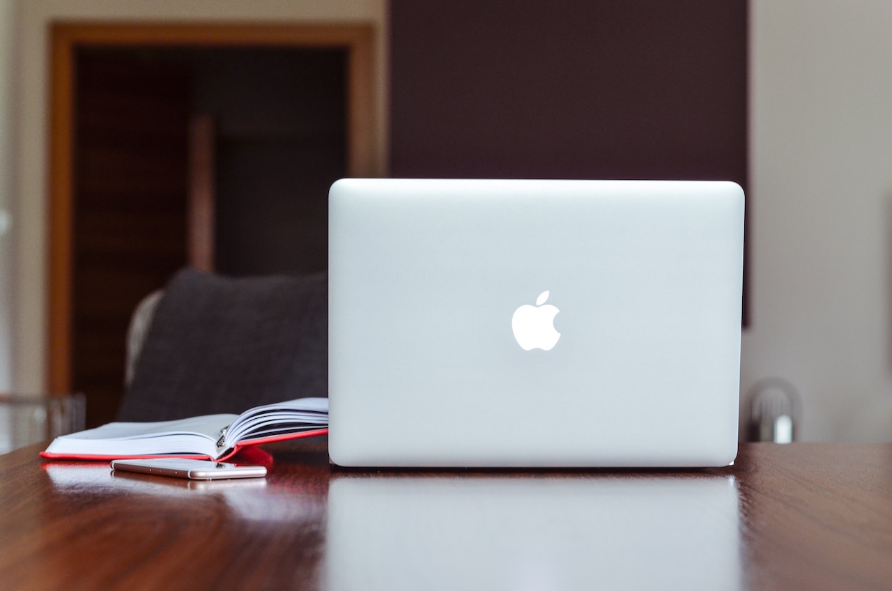 En moderne MacBook med Retina-skjerm står åpen på en minimalistisk arbeidsstasjon, omgitt av en rekke kontorrekvisita som en pennholder, tastatur, og en mus. Bakgrunnen er en nøytral vegg med et kunstverk