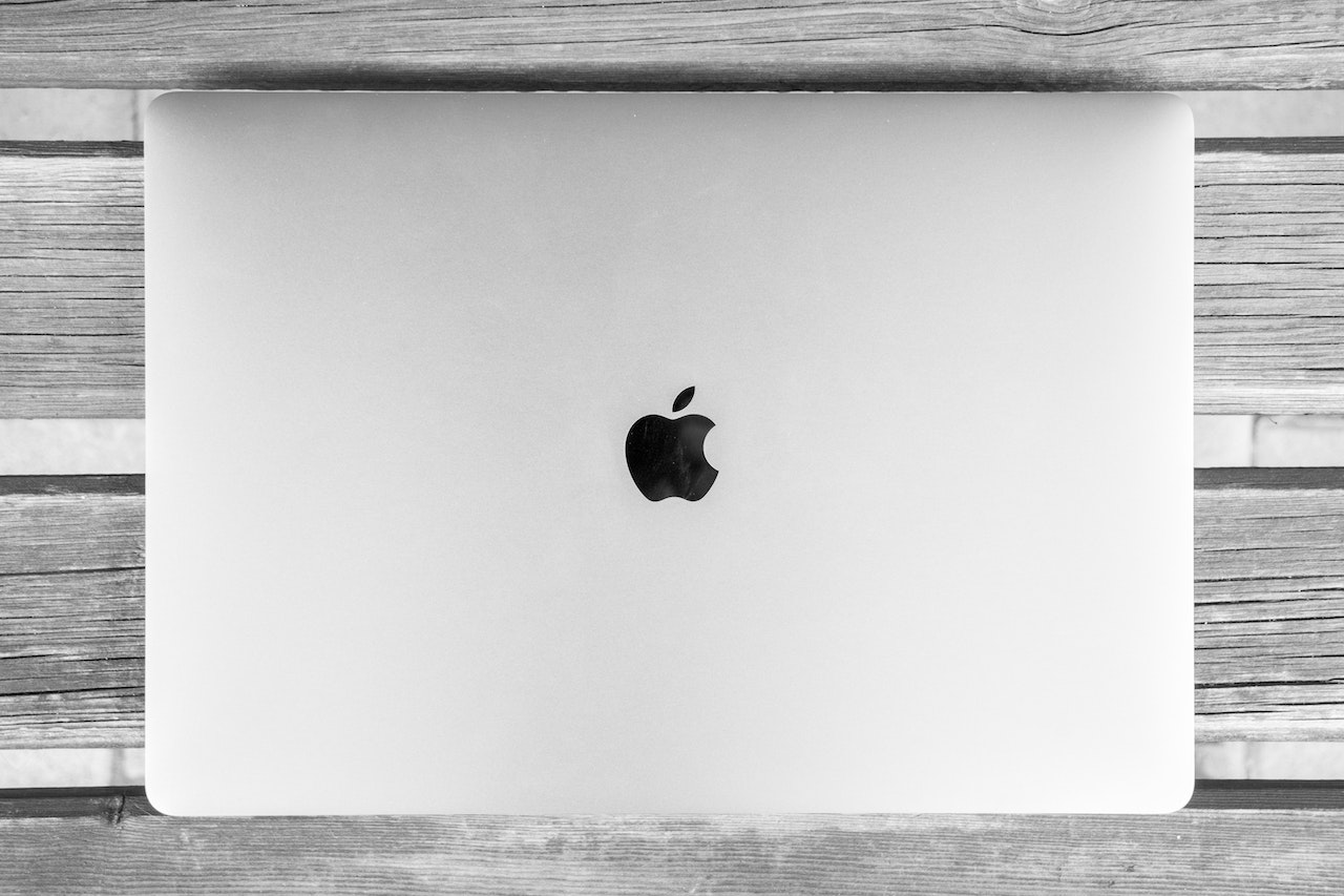 En sølvfarget MacBook med åpen skjerm viser skrivebordet, plassert på et mørkt trebord ved siden av en kopp kaffe og noen notatbøker. Bakgrunnen inkluderer en grønn plante og en vindu