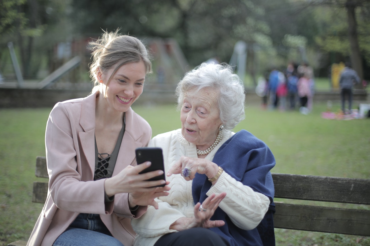 Eldre kvinne sitter ved et bord i et koselig norsk hjem, og lærer seg å bruke en smarttelefon med hjelp fra instruksjonsboken foran seg. Hennes ansiktsuttrykk reflekterer en blanding av konsentrasjon og nysgjerrighet mens hun utforsker den digitale verdenen